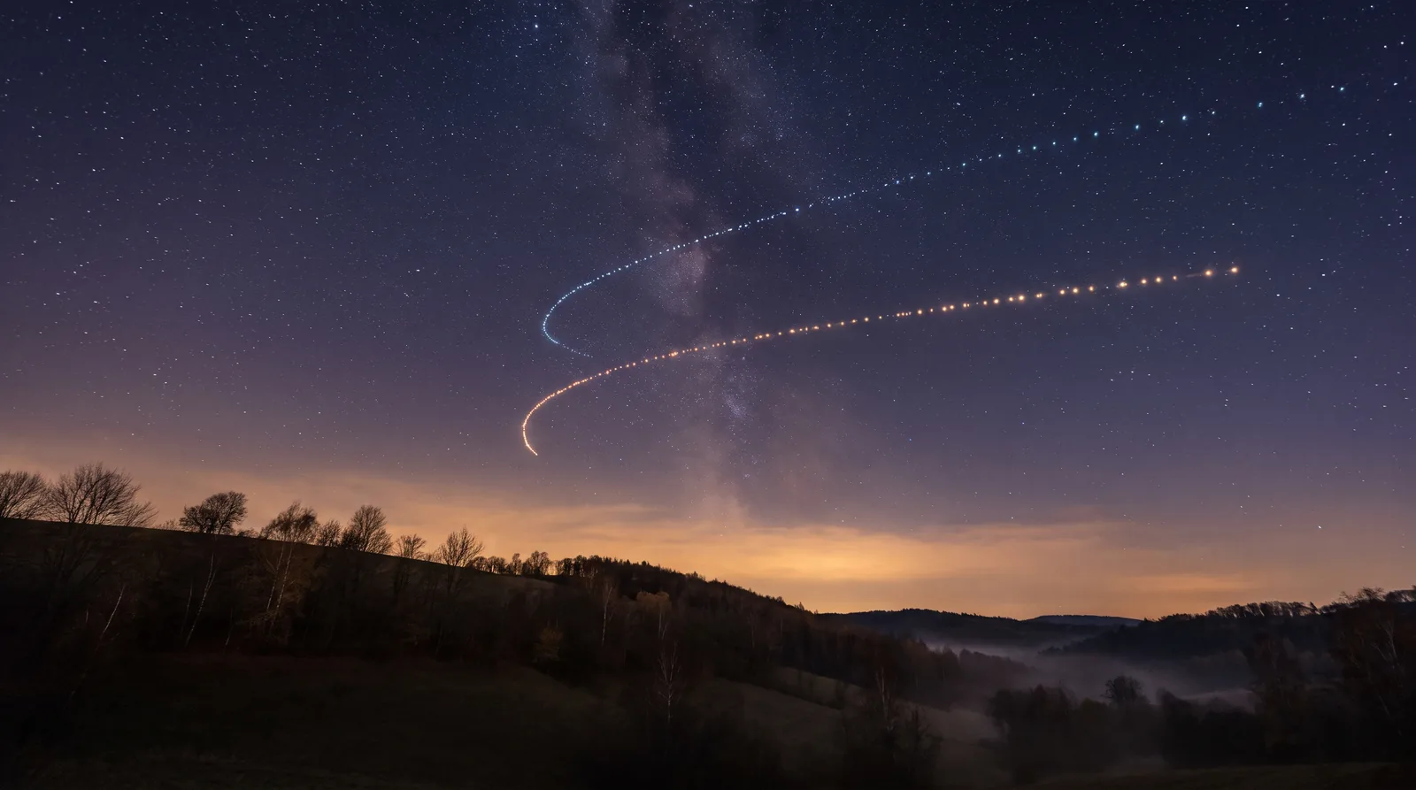 Nachthimmel mit Milchstraße über herbstlichen Hügeln, zwei Planetenbahnen aus Lichtpunkten wenden und laufen wieder nach vorn, im Tal liegt Nebel