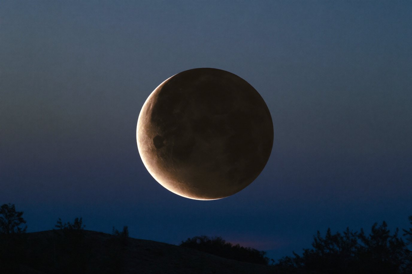 Partiell verfinsterter Vollmond mit schmalem hellem Lichtsaum tief über dunklen Hügeln in der Morgendämmerung